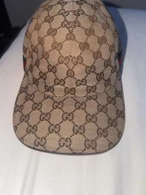  Gucci keps - Säljer en beige keps från Gucci med det klassiska GG-monogrammet över hela kepsen. Kepsen har gröna och röda ränder på sidan och justerbart spänne bak. Tillverkad i canvasmaterial som ger en exklusiv känsla och streetwear-vibe.