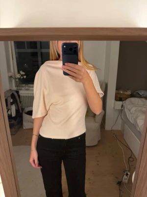 Ljusbeige topp med vid ärm - Säljer en ljusbeige topp med lös passform och en vid, kort ärm. Toppen har en enkel och stilren design med hög halsringning och är tillverkad i ett mjukt material som känns skönt mot huden. Perfekt att styla med jeans eller kjol.