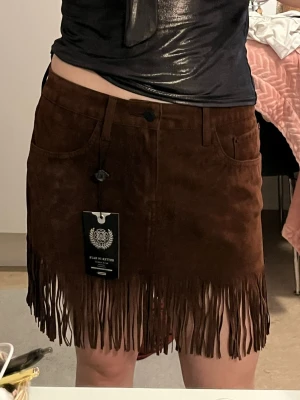 Brun mockakjol med fransar - Säljer en snygg brun minikjol i mockaimitation med fransar längs nederkanten. Kjolen har fickor fram och bälteshällor, vilket ger en cool western-vibe. Perfekt för dig som vill sticka ut med en trendig och unik look. Köpte 600 kr säljer 250 kr.