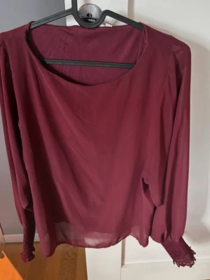 Vinröd blus med långa ärmar - Vinröd blus i tunn, transparent material med rund halsringning och långa ärmar. Ärmsluten har smockdetaljer som ger en snygg och trendig look. Perfekt att matcha med jeans eller kjol för en stilren outfit.