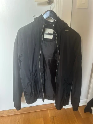 Svart vindjacka med huva - As schysst windbreaker som passar perfekt nu till hösten. Säljs nu för den är för stor och kommer aldrig till användning, pris är ej hugget i sten
