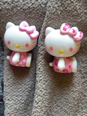 Hello Kitty figurer i rosa klänning - Två söta Hello Kitty figurer i plast med vita kroppar, rosa klänningar och stora rosa rosetter på huvudet. Ena klänningen har prickar och den andra har hjärtan. Perfekta som dekoration eller samlarpryl.