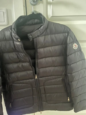 Svart pufferjacka från Moncler - Snygg svart pufferjacka från Moncler med klassisk quiltad design och hög krage. Jackan har dragkedja framtill, två sidofickor med dragkedja och Moncler-logga på ärmen. Perfekt för kalla vinterdagar och riktigt trendig stil.