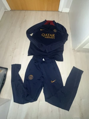 PSG träningsset från Nike, marinblå - Säljer ett PSG träningsset från Nike i marinblått med guldiga detaljer. Setet består av långärmad tröja och byxor, båda med PSG-logga och Nike swoosh. Materialet är Dri-FIT för bra andningsförmåga och komfort. Perfekt för fotbollsträning eller chill.
