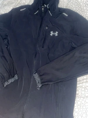 Under Armour windbreaker - Snygg svart vindjacka från Under Armour med dragkedja, huva och reflekterande detaljer. Jackan har logga på bröstet, bröstficka med dragkedja och tryck på ärmsluten. Perfekt för blåsiga dagar och sportiga outfits.