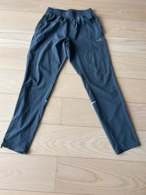 Grå träningsbyxor från Under Armour - Säljer ett par grå träningsbyxor från Under Armour med resår i midjan och fickor på sidorna. Byxorna har smal passform, diskret logga på låret och dragkedjor vid bensluten. Perfekta för löpning eller gym.