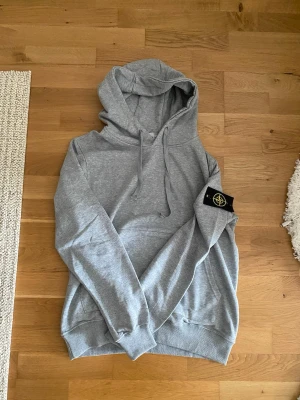 Grå hoodie med Stone Island patch - Snygg grå hoodie från Stone Island med klassisk huva och dragsnören. Hoodien har en ikonisk svart patch med gul logga på ärmen och en stor magficka framtill. Tillverkad i mjukt bomullsmaterial som är perfekt för chill dagar.