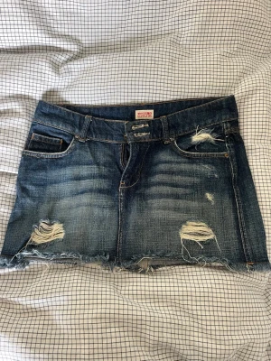 Blå jeanskjol med slitningar - Säljer en kort blå jeanskjol med slitna detaljer och fransig nederkant. Kjolen har klassisk femficksdesign och råa kanter. Jättesnygg kjol, perfekt för sommar eller höst med ett par strumpbyxor under! 