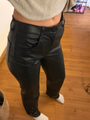Svarta raka jeans i fuskskinn - Säljer ett par svarta jeans i fuskskinn med hög midja och raka ben. Byxorna har framfickor och stängs med knapp och dragkedja. Perfekta för en trendig och edgy stil.