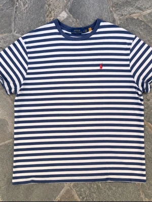 Randig t-shirt från Polo Ralph Lauren - Säljer en klassisk blåvit randig t-shirt från Polo Ralph Lauren med röd broderad logga på bröstet. T-shirten har korta ärmar, rund halsringning och är gjord i mjuk bomull. Perfekt för en avslappnad och stilren look.