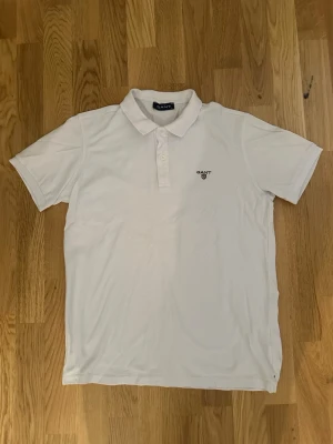 Gant pikétröja  - Klassisk vit pikétröja från Gant med korta ärmar och krage. Tröjan har knäppning med tre knappar framtill och en liten broderad Gant-logga på bröstet. Perfekt för en stilren och avslappnad look.