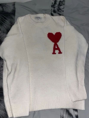 Vit stickad tröja från Ami - Vit stickad tröja från Ami Alexandre Mattiussi med rund halsringning och ribbade muddar. På bröstet finns ett rött hjärta och bokstaven A. Perfekt för dig som gillar stilrena och trendiga plagg.
