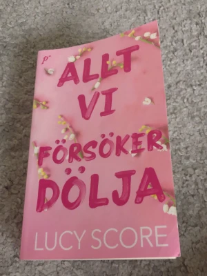 Allt vi försöker dölja av Lucy Score - En romantisk och humoristisk roman med rosa omslag och vita liljekonvaljer. Boken handlar om polischefen Nash Morgan och Lina Solavita, fylld av hemligheter, vänskap och passion. Perfekt för dig som gillar småstadskänsla och starka karaktärer.