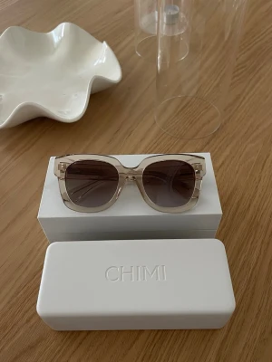 Beige solglasögon från CHIMI - Snygga solglasögon från CHIMI med transparent beige båge och fyrkantig form. Glasen är mörka och ger en cool kontrast till den ljusa bågen. Kommer med originaletui. Perfekta för dig som vill ha en stilren och modern look.