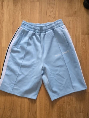 Palm Angels shorts ljusblå  - Snygga ljusblå shorts från Palm Angels med vita sidoränder. Modellen har elastisk midja och två fickor framtill. Perfekta för en avslappnad och trendig stil. 