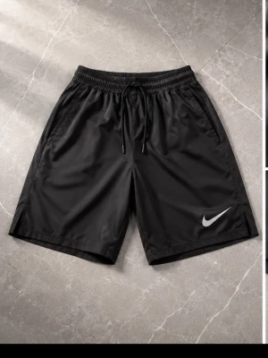 Svarta träningsshorts från Nike - Svarta träningsshorts från Nike med elastisk midja och snörning. Shortsen har en enkel och sportig design med Nike-logga på benet. Tillverkade i ett lätt och snabbtorkande material som passar perfekt för träning eller varma dagar.