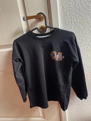 Svart sweatshirt Los Angeles ALAA - Cool svart sweatshirt från ALAA med tryck 'Los Angeles' och stora bokstäver på bröstet. Tröjan har rund halsringning och ribbade muddar vid ärmslut och nederkant. Perfekt för en avslappnad och trendig look.