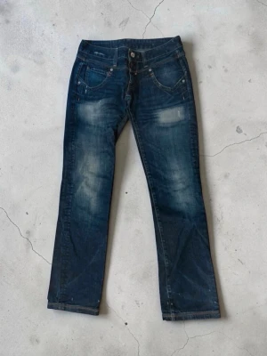 Mörkblå bootcut jeans med slitningar från replay  - Snygga mörkblå jeans med bootcut passform och slitna detaljer framtill. Jeansen har klassiska fem fickor, dubbla knappar i midjan och ljusa tvättade partier för en cool look. Perfekta för dig som gillar en avslappnad men trendig stil.