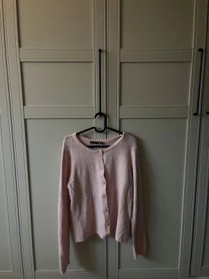 Ljusrosa stickad kofta - Söt ljusrosa stickad kofta med rund halsringning och knappar framtill. Koftan har långa ärmar och är gjord i ett mjukt, fluffigt material som känns skönt mot huden. Perfekt för en mysig look. Lite nopprig, från lager 157