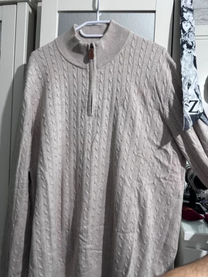 Beige kabelstickad half-zip Redford XL - Snygg beige kabelstickad tröja från Redford i storlek XL. Tröjan har halv dragkedja med ribbad hög krage och diskret logga på bröstet. Perfekt för lager på lager och passar till jeans eller chinos. Materialet känns mjukt och bekvämt.