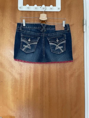 Mörk jeanskjol med spetskant - Snygg mörkblå jeanskjol med dekorativa bakfickor och kontrastsömmar. Kjolen har en rosa spetskant längst ner som ger en extra detalj. Klassisk femficksmodell med knapp och dragkedja framtill. Pris kan förhandlas och skickar fler bilder vid förfrågan.