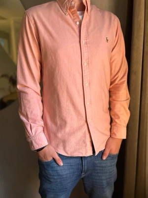 Ralph Lauren Skjorta Ljus Rosa - Säljer en stilren ljusrosa skjorta med klassisk krage och knäppning framtill. Skjortan har långa ärmar och en liten broderad logga på bröstet. Perfekt för dig som vill ha en enkel men snygg skjorta till jeans eller chinos. Ralph Lauren Skjorta