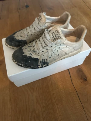Maison Margiela paint splatter sneakers - Unika sneakers från Maison Margiela i beige canvas med svart och grå paint splatter-design. Klassisk låg modell med vita snören och synlig etikett på plösen. Snygg kontrast mellan den ljusa basen och de mörka färgstänken. Perfekta för dig som gillar statement-plagg.