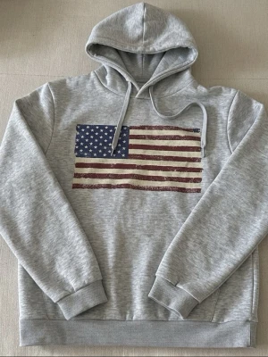 Grå hoodie med amerikansk flagga - Säljer en grå hoodie med tryck av amerikanska flaggan på bröstet. Hoodien har huva med dragsnören och en stor magficka framtill. Perfekt för dig som gillar en avslappnad och stilren look. Skriv för fler frågor!!