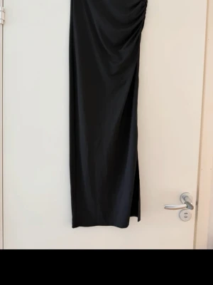 Svart långkjol med slits - Säljer en svart långkjol i ett glansigt och mjukt material. Kjolen har en hög slits på sidan som ger en snygg och trendig look. Passar perfekt till dig som gillar minimalistisk stil med en twist. Skulle säg att den är som en vanlig xs