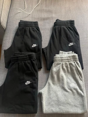 Nike mjukisbyxor  - Fyra par mjukisbyxor från Nike i svart och ljusgrått. Klassisk design med resår i midjan och muddar vid benslut. Byxorna har sidofickor och Nike-logga broderad på vänster ben. Perfekta för chill eller träning. Stl 158-170 (Youth XL). Ignorera den andra storleken som är angiven.