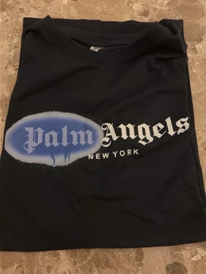 Svart Palm Angels t-shirt med tryck - Säljer en svart t-shirt med stort Palm Angels-tryck i vitt och blått på bröstet samt texten 'New York' under. Klassisk rund hals och korta ärmar. Tillverkad i mjuk bomull, perfekt för en avslappnad streetwear-look.