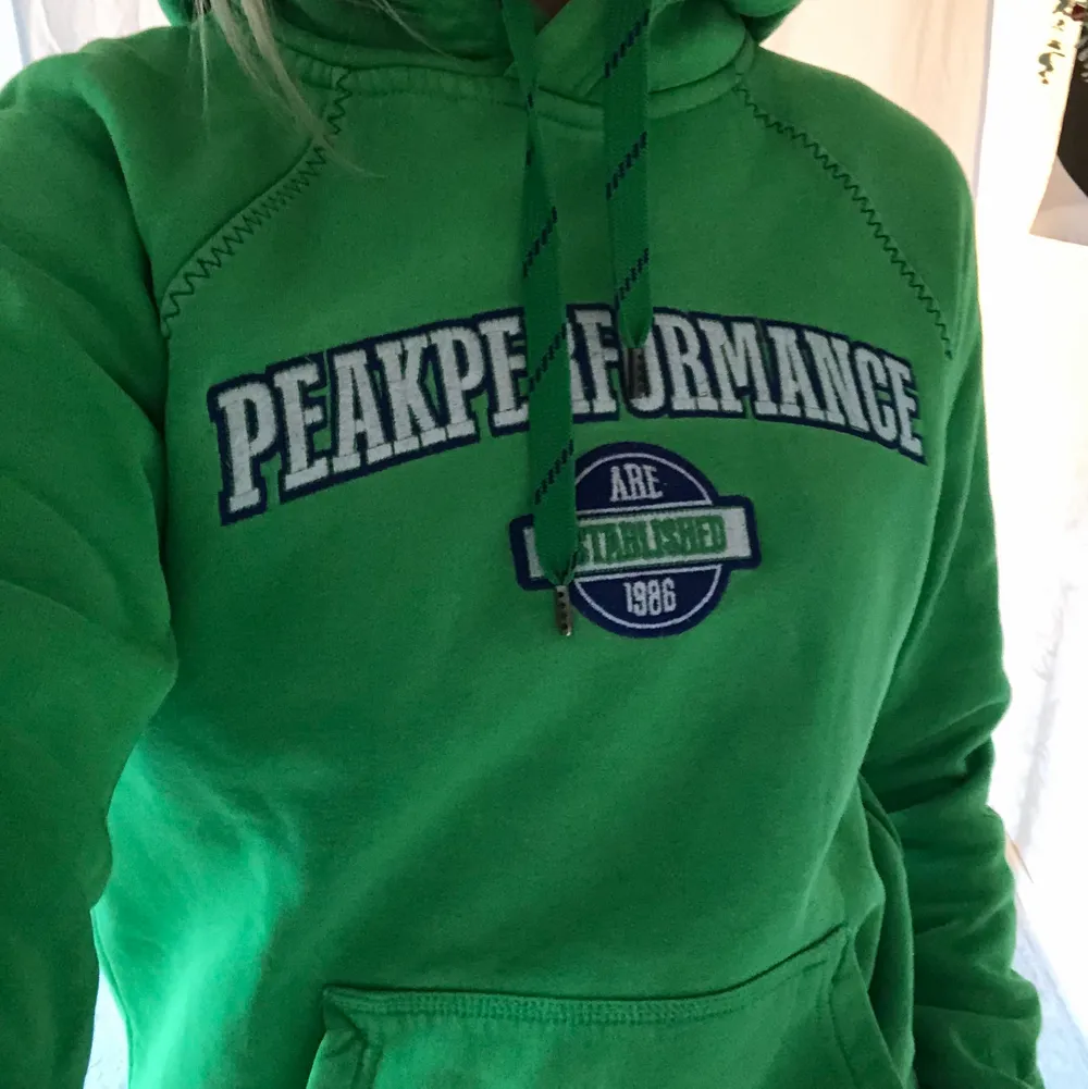 En ljus grön peakperformance hoodie (luvtröja) har detaljer med vit och blått på märket. Luvan har två snören som syns där fram på tröjan.. Tröjor & Koftor.