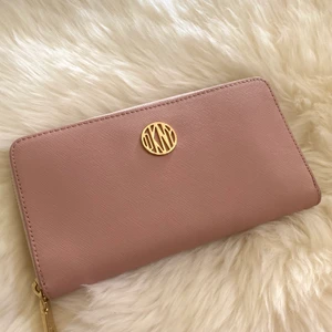 DKNY plånbok  - Plånbok från DKNY. Köpt för 1200 på Åhléns. Rosa med guld detaljer. 8 fack för kort samt 3 fack för kontanter, en med dragkedja för mynt i mitten. Finns att hämta i Alby eller kan mötas upp i Stockholms området. Kan skickas i posten (köparen står för frakten)