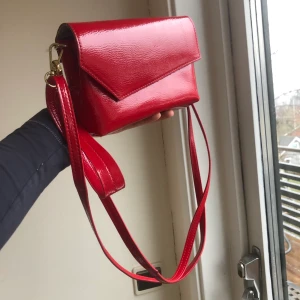 Röd väska - Röd liten väska i glansigt material. Kommer med ett längre axelband som man kan justera och ett litet band för att hålla i handen. Ny skick