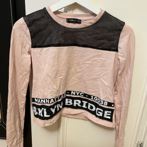 Croppad babyrosa långärmad - En ljusrosa Croppad långärmad med svart tryck och vit text samt ett svart nät på fram-och baksida. Inhandlad på Newyorker