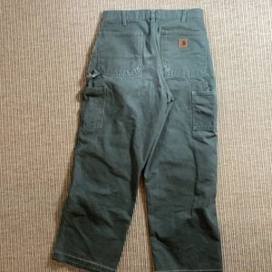 Carhartt carpenter byxa  - Olivgröna carhartt single knee carpenter pants. Det är vintage och är lite slitna och har på högersida en lagning. Det är en ”released hem” längst ner. Enkelt att det fixa ifall man vill få tbx till orginal längd.                W30xL30. I midjan 39cm, längd 103cm. Kan mötas upp och frakt ingår i priset. 