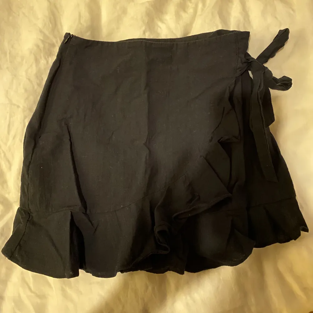 Supersöt svart omlottkjol med volang och knytdetalj på sidan. Kjolar.