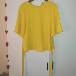 Gul blus med vida ärmar - Plagget har en liten mascara fläck på insidan av kragen som jag ite lyckats få bort men den syns inte när du har den på dig😌 blusen är knytbar med fastsydda band på var sida samt en slits på frasidan ovanför bysten. Valt pris plus frakt på 49kr