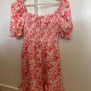 Blommig klänninh - Klänning i färgerna rosa/ röd aktiga färger.  Klänningen har ett prydnadsband över bröstet samt super härliga puffärmar. Nypris 130 Mitt pris 70