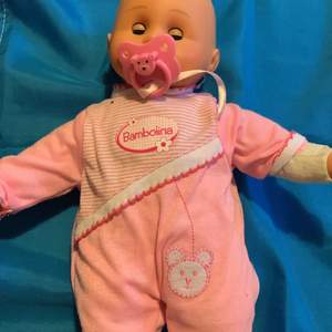 Det är en bebis med en rosa overall. Den har också en rosa napp med björnform. 