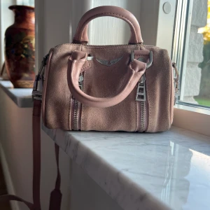 Liten ljusrosa, mocka Zadig väska  - Säljer denna ljusrosa mocka Zadig Sunny nano bag. Jättefin och väldigt rymlig för att vara så liten. Nypris cirka 1800kr FRAKTEN BETALAR KÖPAREN!! Väskan än tyvärr ganska smutsig Säljer den för jag inte gillar den lika mycket som när jag köpte den