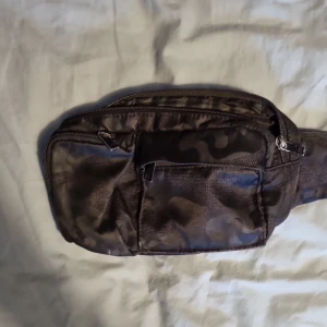 Midjeväska - Säljer denna svarta midjeväskan med militärmönster. Ett litet hål vid ena dragkedjan men den går fof. att dra igen. Dragtapperna/handtagen är slitna i färgen. Den går att justera och knäppa upp, lite smutsig längst ner men går o tvätta bort. Kontakta