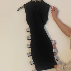 Klubbklänning - En mini svart klänning med öppning på vardera sida. Klänningen sitter bra på kroppen, har stretch och framhäver kroppen och speciellt med extra detaljern  på sidorna. Finns dragkedja på ryggen av klänningen. Klänningen har använts enbart en gång. 