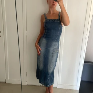 Jeansklänning från Socex - En stilren jeansklänning från Socex i storlek 34. Klänningen har justerbara axelband och en rak passform. Den är ärmlös och har en medellång längd som sträcker sig till knäna.