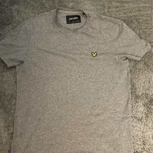 Grå t-shirt från Lyle & Scott - En grå t-shirt från Lyle & Scott med en liten gul logotyp på bröstet. T-shirten har korta ärmar och en rund halsringning. Passar L och M