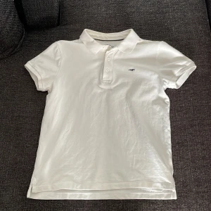 Vit pikétröja från Hampton Republic - Säljer en snygg vit pikétröja från Hampton Republic i storlek 146/152. Tröjan är i bra skick och har korta ärmar samt en klassisk krage med knappar. Den är gjord av bomull och har en liten broderad logga på bröstet. Perfekt för sommarens alla aktiviteter! 🌞