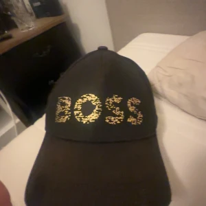 Svart keps från BOSS - Säljer en snygg svart keps från BOSS med guldglittrande text på framsidan. Kepsen är i nyskick och har en justerbar rem på baksidan för perfekt passform. Perfekt accessoar för både vardag och fest!