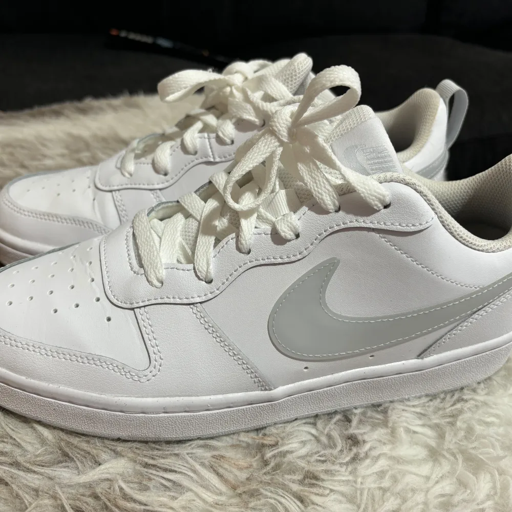 Säljer ett par vita Nike sneakers i nyskick. Skorna har en klassisk design med en vit ovandel och en grå Nike-logga på sidan. De är tillverkade i syntetmaterial och har snörning framtill. Perfekta för vardagsbruk eller träning.. Skor.