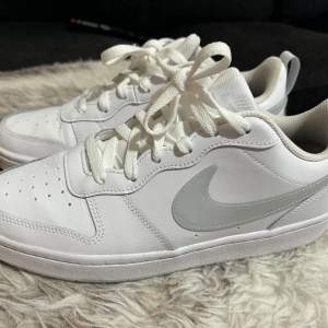 Säljer ett par vita Nike sneakers i nyskick. Skorna har en klassisk design med en vit ovandel och en grå Nike-logga på sidan. De är tillverkade i syntetmaterial och har snörning framtill. Perfekta för vardagsbruk eller träning.