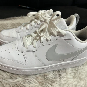 Vita Nike sneakers - Säljer ett par vita Nike sneakers i nyskick. Skorna har en klassisk design med en vit ovandel och en grå Nike-logga på sidan. De är tillverkade i syntetmaterial och har snörning framtill. Perfekta för vardagsbruk eller träning.